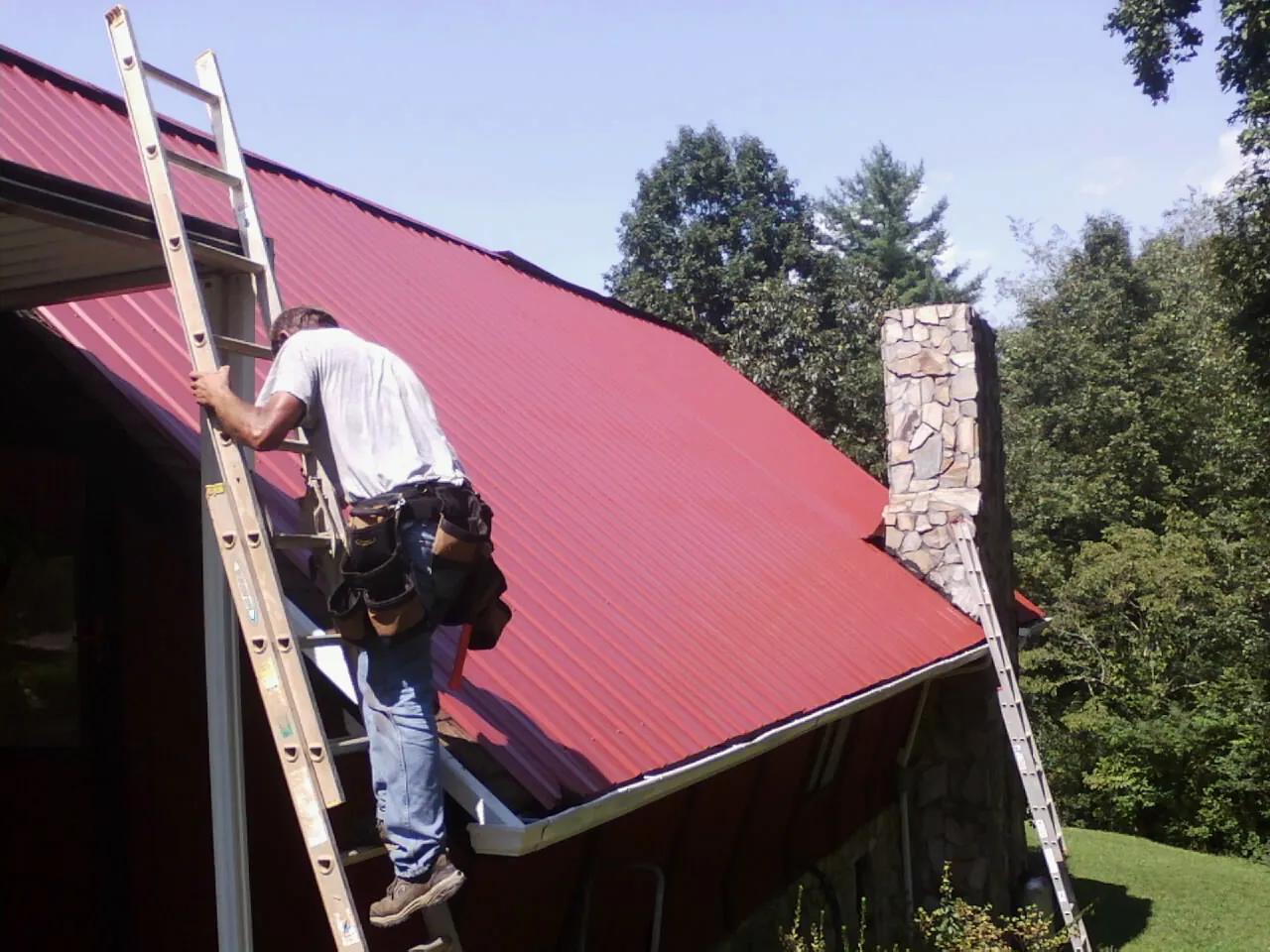 Roofer on a ladder beside a red metal roof and stone chimney — Soffit & Fascia Repair by Roofing Brothers in David City
