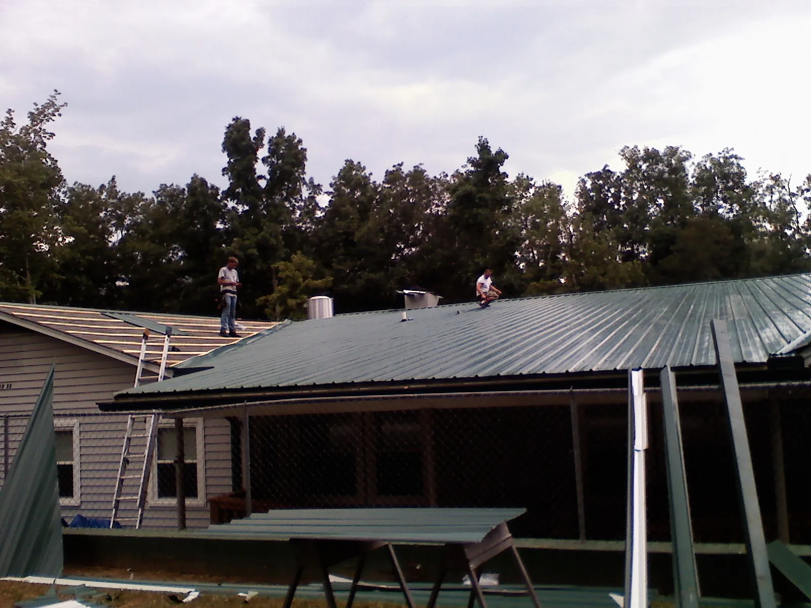 Two roofers installing green metal panels above a covered porch in David City